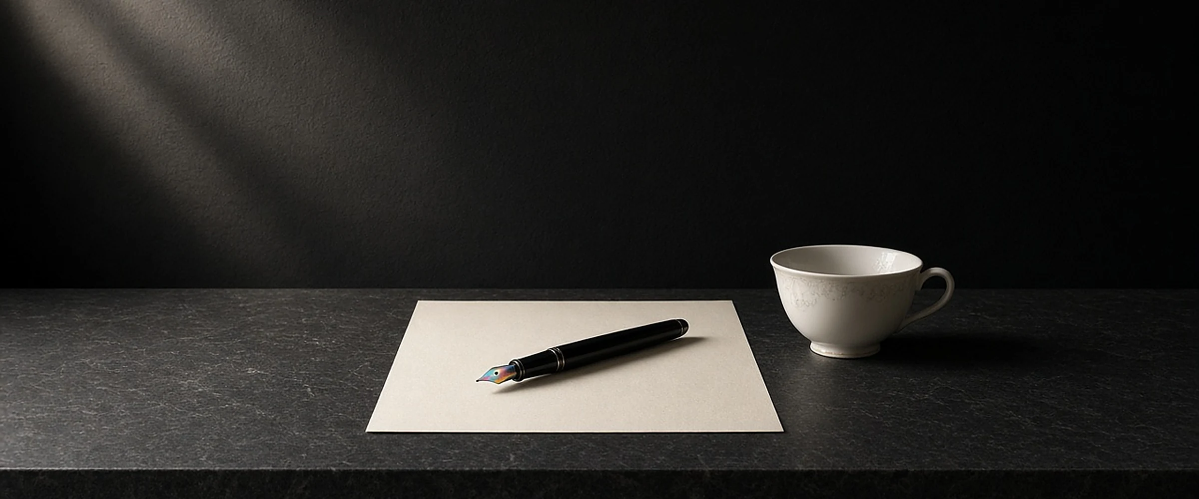 A fountain pen resting on a blank sheet of paper beside a porcelain teacup on a dark stone ledge.