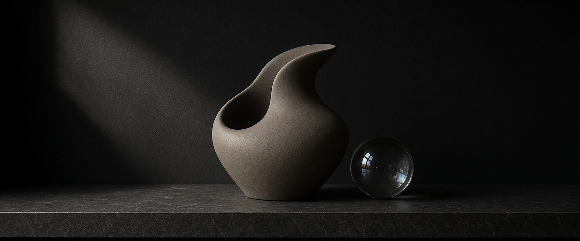 A sculptural matte ceramic vase and a glass sphere on a dark ledge in a beam of light.