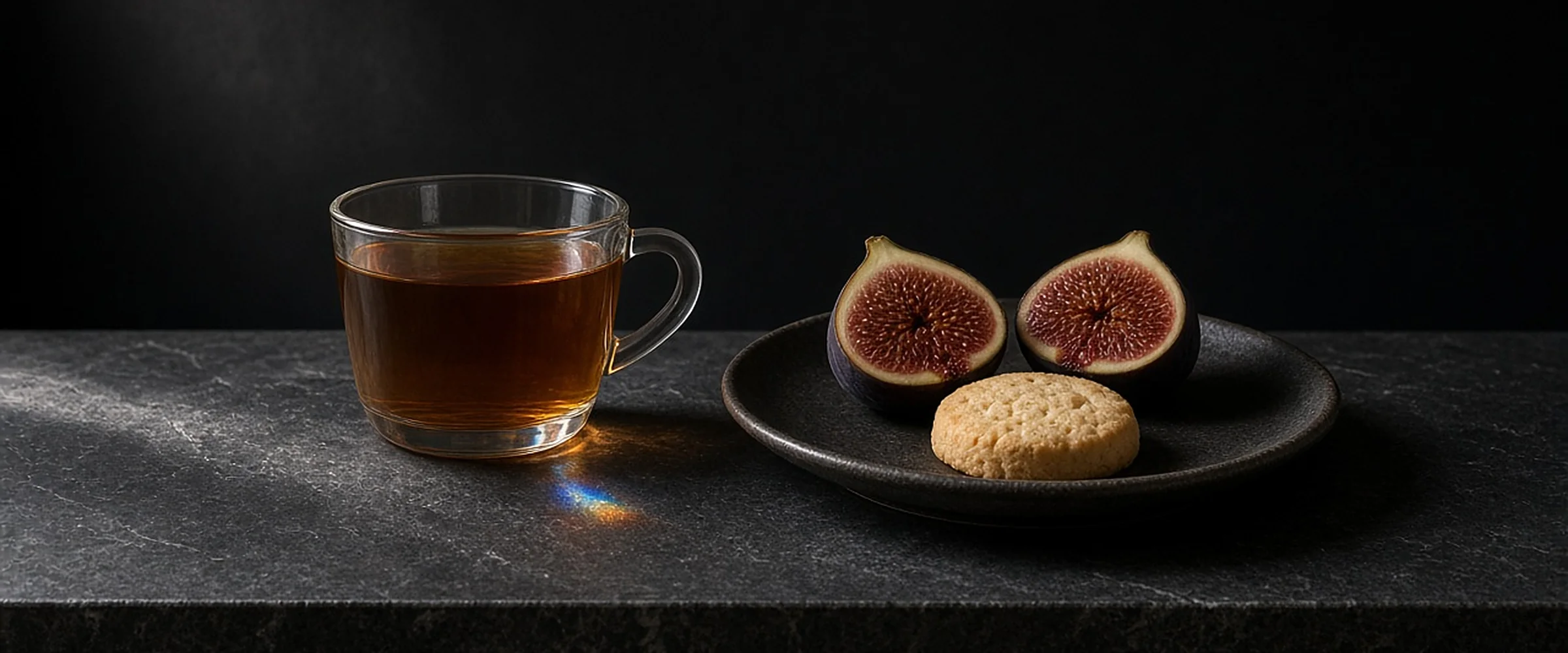 A glass cup of amber tea beside halved figs and a shortbread biscuit on a dark plate.