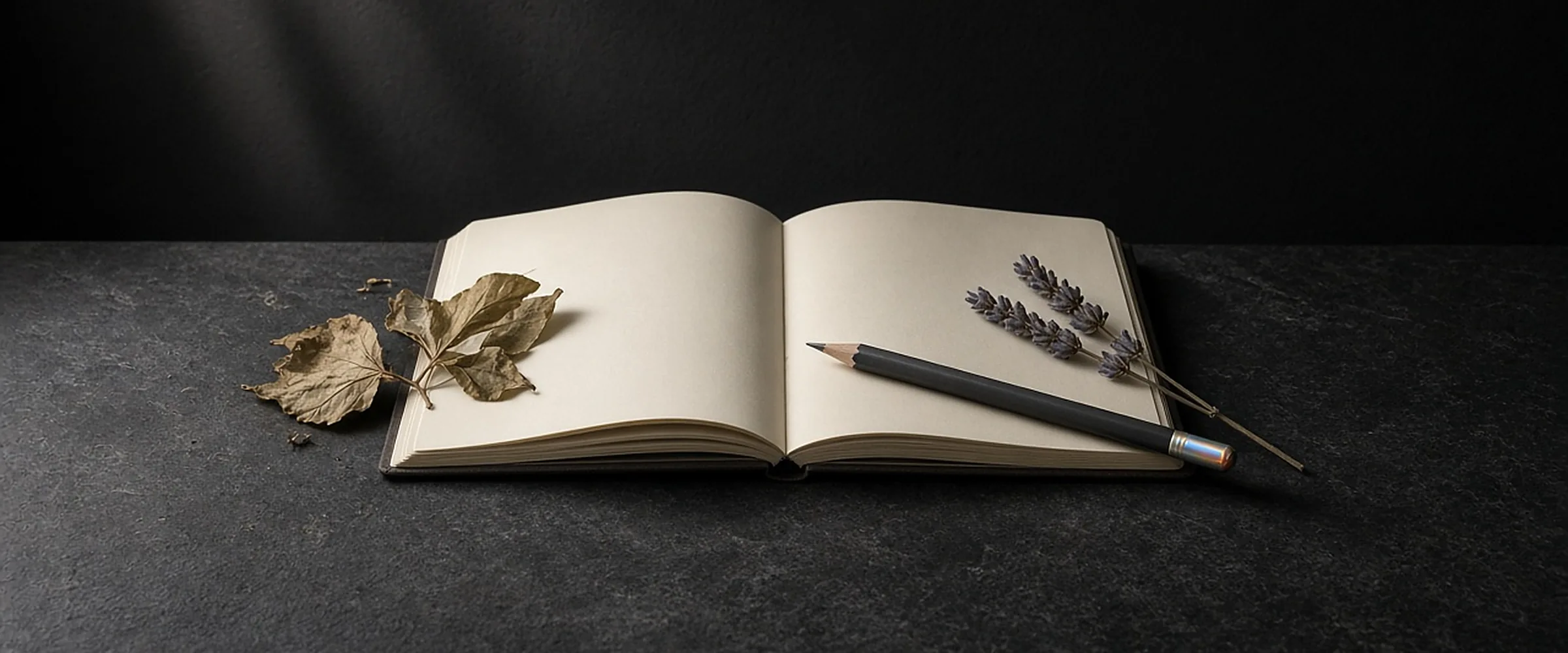 An open blank notebook with a pencil, pressed leaves and dried lavender on dark stone.