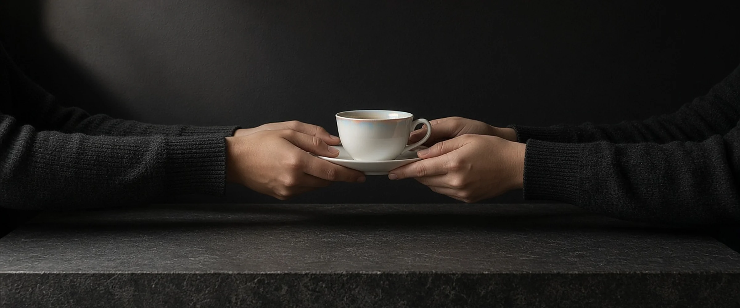 Two pairs of hands passing a porcelain teacup on a saucer across a dark stone ledge.