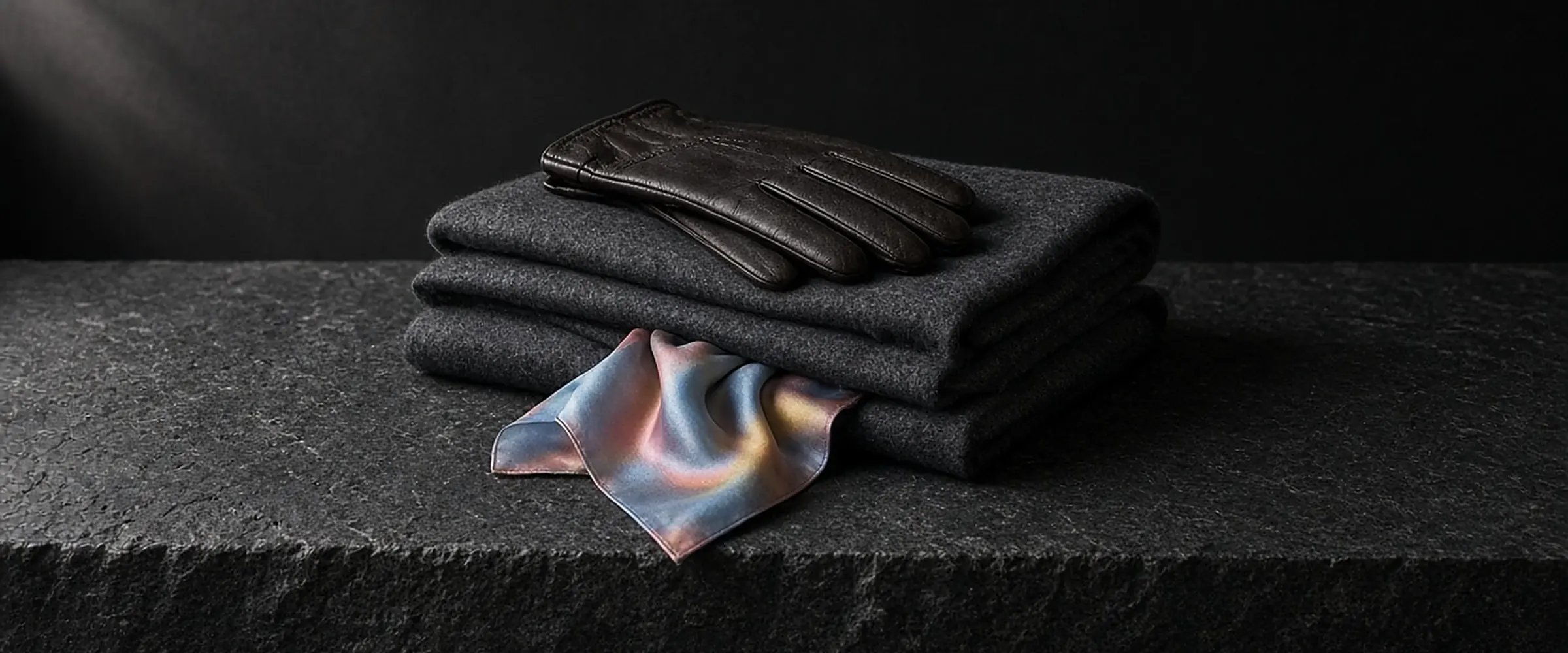 Leather gloves on a folded charcoal wool scarf, with an iridescent silk pocket square, on dark stone.